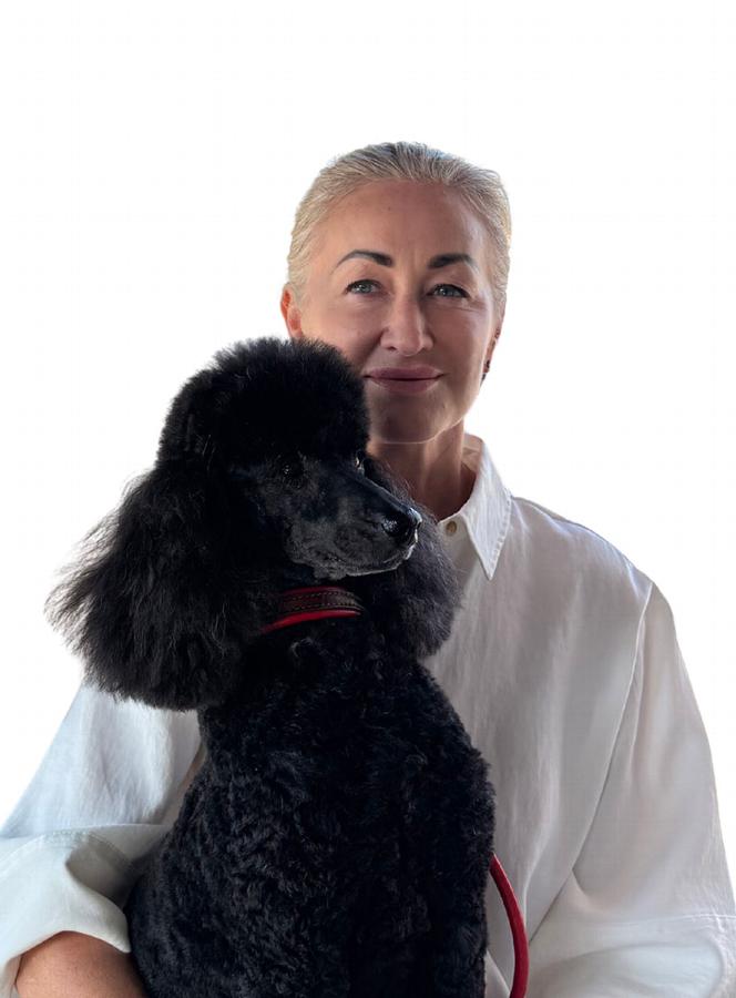 A woman holding a black poodle, both smiling, with a light background.